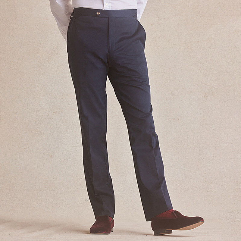 A man standing wearing navy flat front cotton chinos with a white shirt and red   velvet slippers.